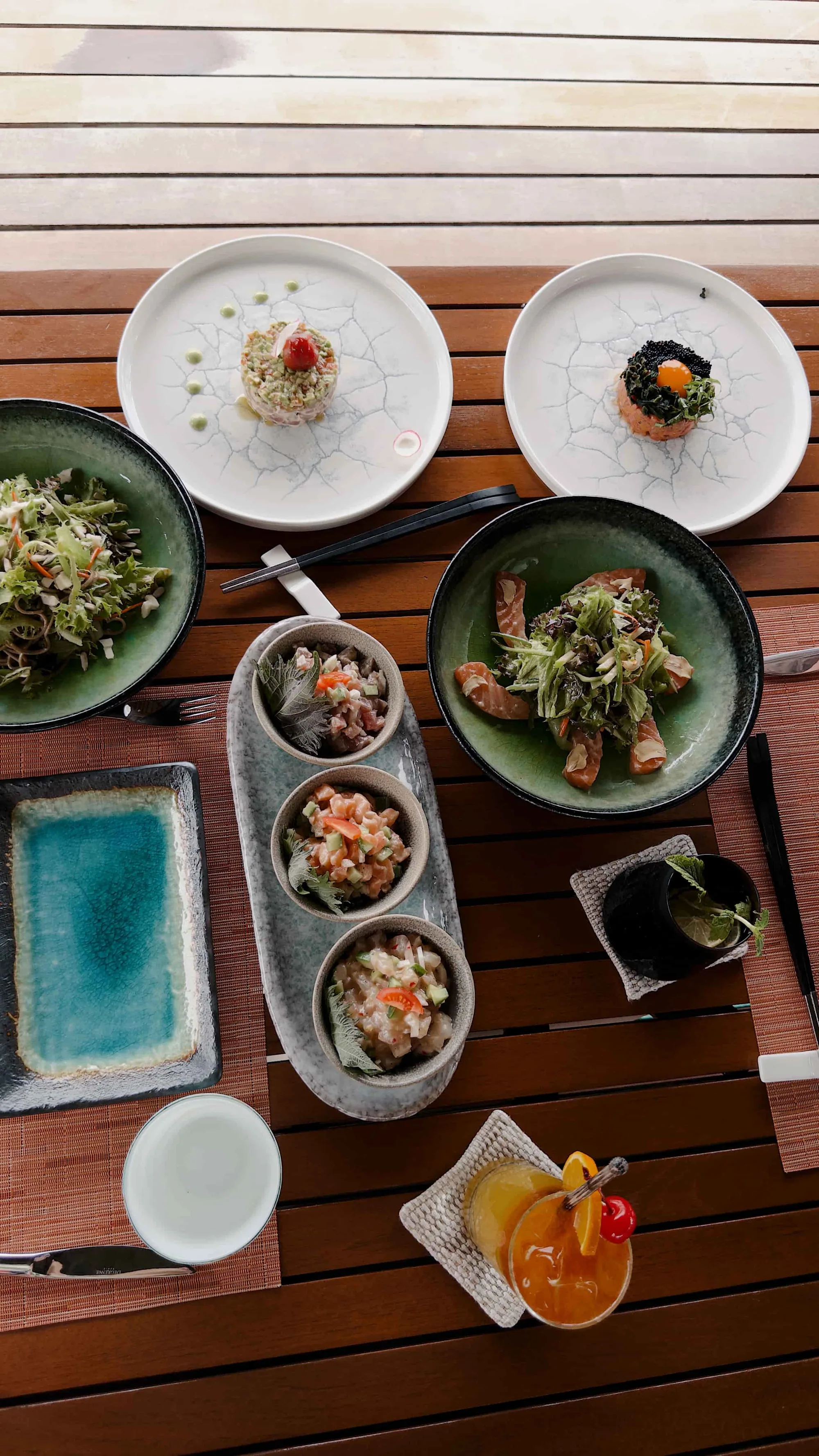 Huvafen Fushi, dining, North Malé Atoll, Maldives, SALT izakaya plate spread, top-down composition on dark timber decking: two web-glaze tartare plates, two earthen bowls with greens and grilled protein, trio of ceviche cups in a stoneware holder, sake and a citrus cocktail.