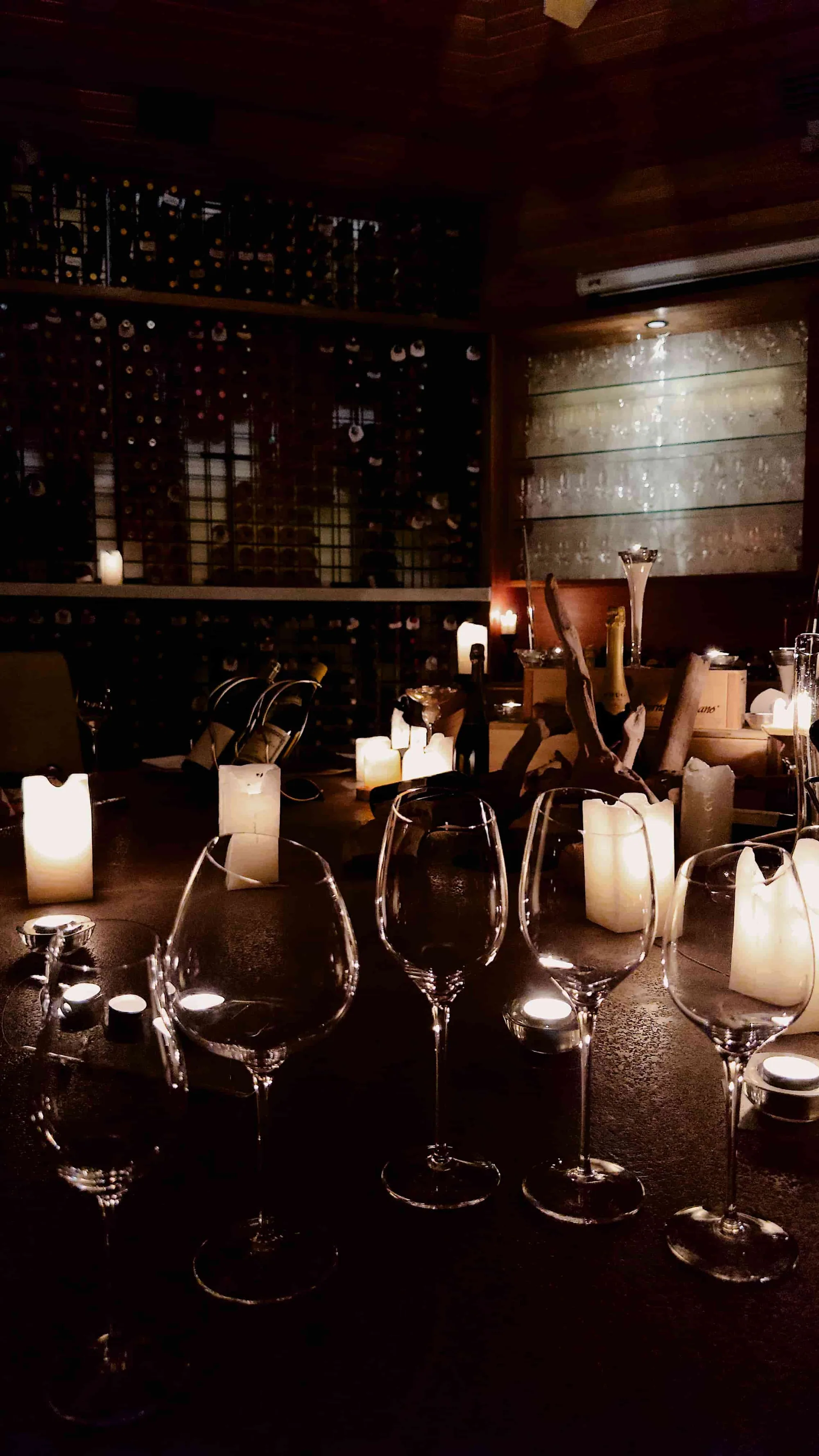Huvafen Fushi, dining, North Malé Atoll, Maldives, Vinum tasting moment, four crystal wine glasses on the stone counter with pillar candles and an open champagne bottle in the chiller, the bottle wall and backlit glassware reading in the warm cellar light.