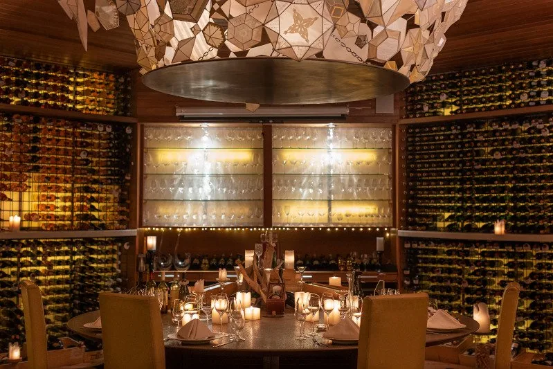Huvafen Fushi, dining, North Malé Atoll, Maldives, Vinum subterranean cellar wide view, the property's signature origami-style sculptural ceiling above a round dining table, curved walls of dark-timber wine racks with backlit glassware shelves and candle-lit table setting.