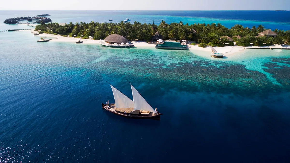 Huvafen Fushi, aerial, North Malé Atoll, Maldives, Traditional dhoni sailing past the boutique-scale island, white sails set against the deep-water channel with the arrival jetty, infinity pool pavilion and palm-fringed villa cluster behind, overwater villas extending left along the reef.
