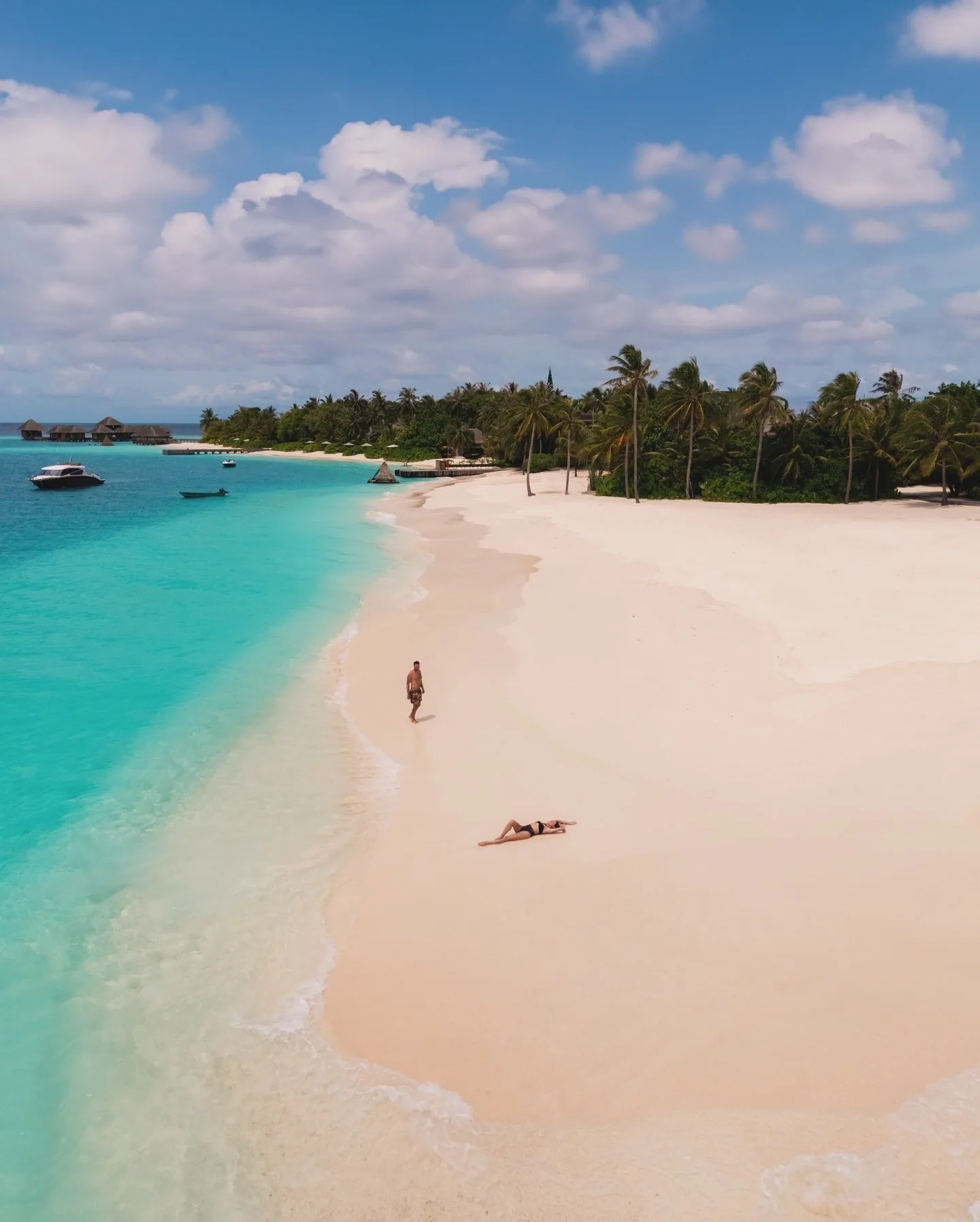 Huvafen Fushi, setting, North Malé Atoll, Maldives, Wide drone view of the resort beach, the white-sand crescent edged by the palm-dense interior, two figures on the shoreline for scale, dhoni and speedboat moored against the LIME spa pavilion cluster visible on the far left.