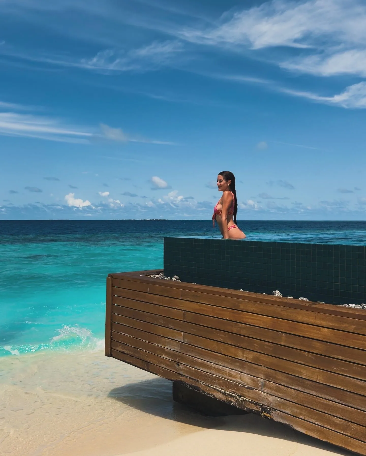 Huvafen Fushi, setting, North Malé Atoll, Maldives, Main pool cantilever-edge detail from the beach side, weathered-timber cladding wrapping the infinity-pool platform with the deep-blue lagoon falling away below, a guest standing at the dark-tile pool edge for scale, the white-sand crescent and reef shelf visible left.