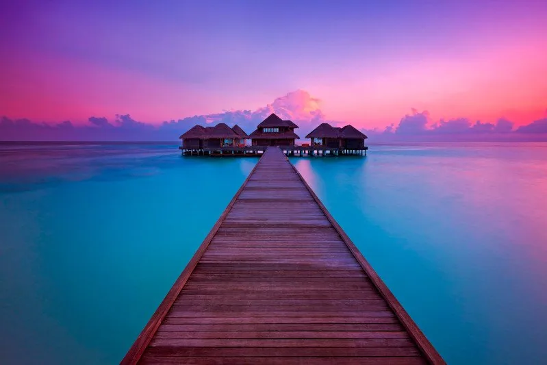 Huvafen Fushi, spa, North Malé Atoll, Maldives, LIME spa pavilion cluster at twilight, the long timber jetty leading out to the silhouetted thatched treatment pavilions, pink-and-violet sky reflecting on the still lagoon.