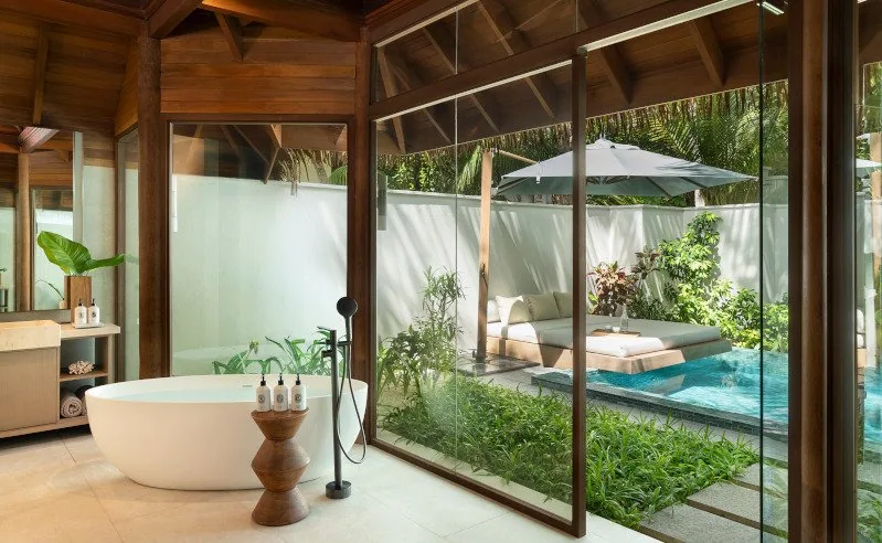 Huvafen Fushi, villa beach, North Malé Atoll, Maldives, Beach Pavilion bathroom, free-standing oval tub with matte-black floor mount, dark-timber rafters under a pitched ceiling, full-height glass walls opening onto the private courtyard daybed under a canvas umbrella and the plunge pool framed by lush planting.