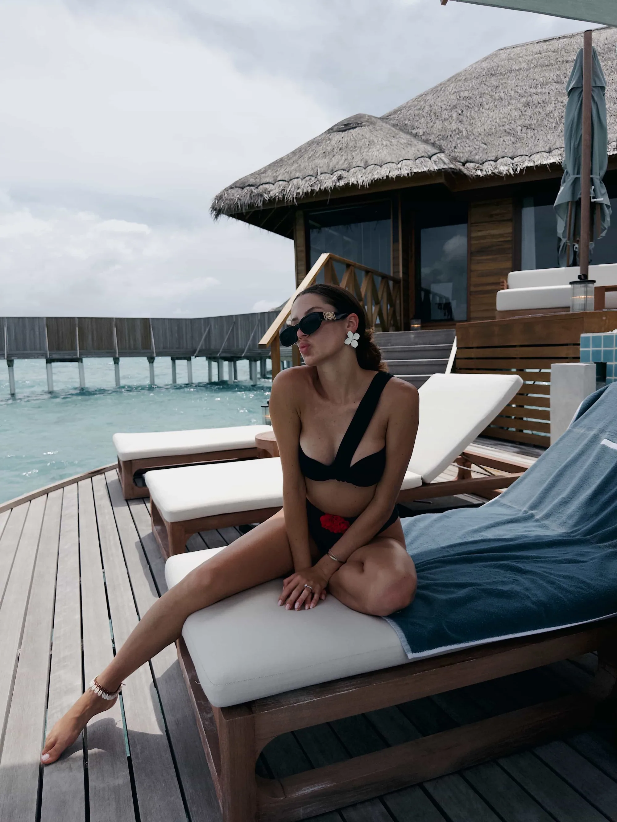 Huvafen Fushi, villa overwater, North Malé Atoll, Maldives, Overwater Pavilion sun deck, timber slatted pergola above a daybed dressed with magenta-striped cushions, woven-rattan privacy screen, a guest leaning into the lagoon-edge plunge pool with the thatched roof of the next pavilion visible against the open horizon.