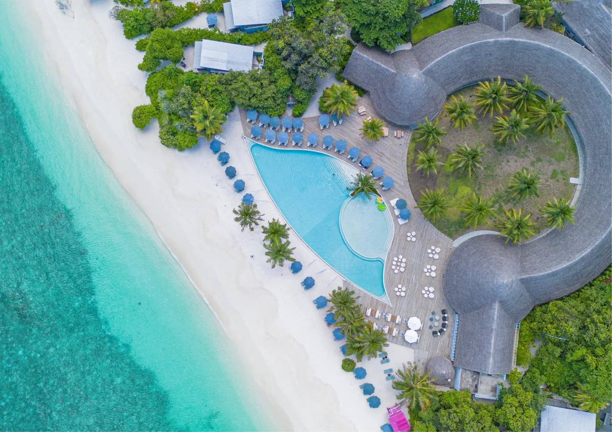 Ifuru Island Maldives, aerial, Raa Atoll, Maldives, Main-pool drone aerial showing the dumbbell-shaped pool with a circular thatched-roof central pavilion at one end, palm-shaded sun-lounger rows in blue parasols, and the white-sand beach edge of the 1.2-kilometre west-facing frontage.