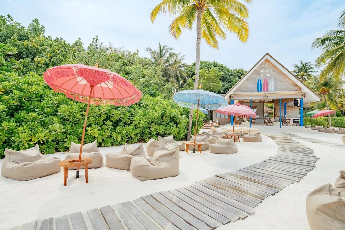 Ifuru Island Maldives, dining, Raa Atoll, Maldives, The Beach Club casual day venue with the painted clapboard pavilion (surfboard signage above the entrance), pink and blue tasseled umbrellas over sand-set beanbag clusters, and the plank boardwalk weaving through the seating arrangement.