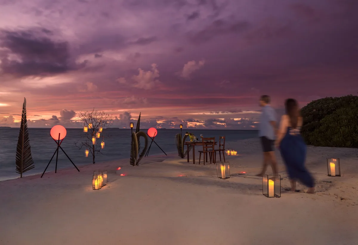 Ifuru Island Maldives, dining, Raa Atoll, Maldives, Hubba Hubba sundowner destination-dining setup with hurricane-glass lantern path receding into the dusk lagoon horizon, sculptural palm-frond candleholders, illuminated red orb lights, a couple walking the beach in dusk silhouette along the 1.2-kilometre west-facing axis.
