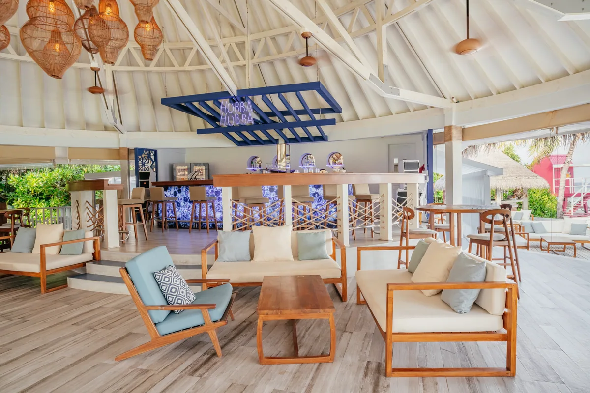 Ifuru Island Maldives, dining, Raa Atoll, Maldives, Hubba Hubba bar interior under the pavilion roof with woven pendant lights, blue stylised signage, white columns, blue-and-white floral upholstered bar chairs at the counter, teak sofa-and-coffee-table lounge groupings, the pink sundowner tower visible through the side opening.