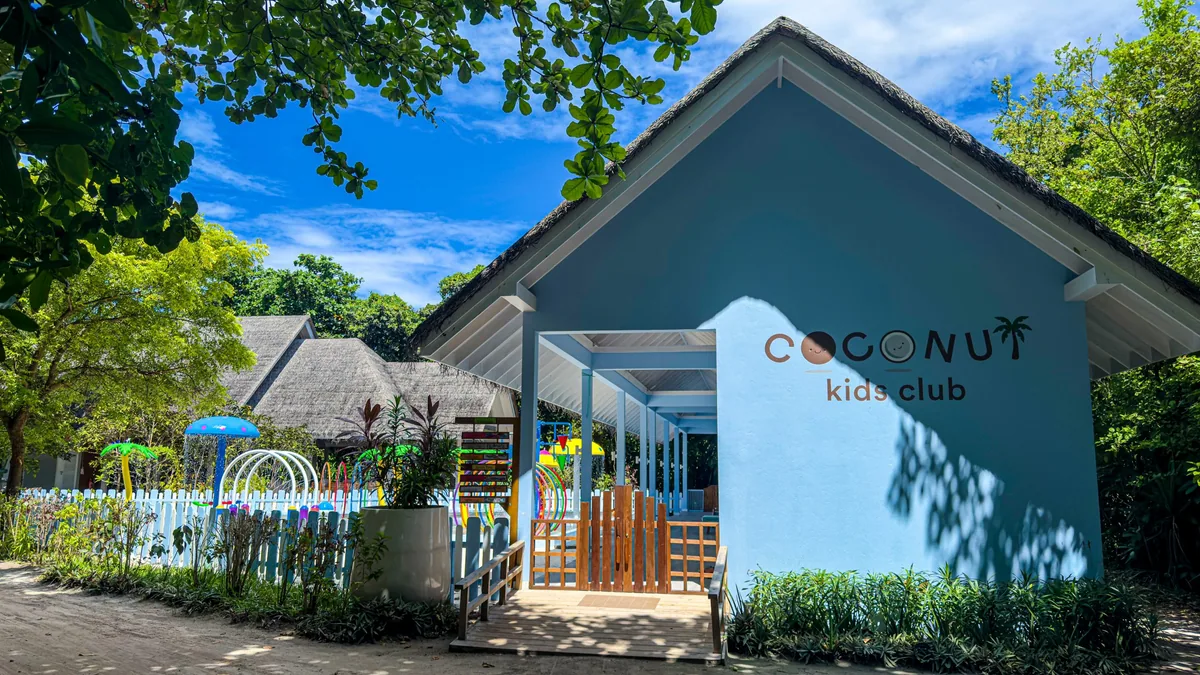 Ifuru Island Maldives, lifestyle, Raa Atoll, Maldives, Coconut Kids Club branded pavilion (pale-blue gabled thatched roof with palm-icon wordmark), white picket fence enclosing the splash-zone water park with green, red, and blue spray toys.