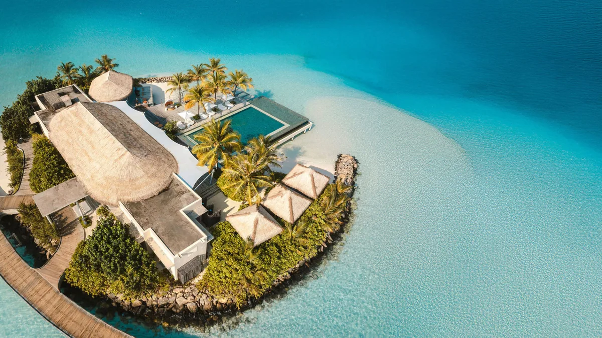 InterContinental Maldives Maamunagau Resort, aerial, Raa Atoll, Maldives, DJI aerial of the small connecting islet between the two sides of Maamunagau Island, showing the elevated central pool deck with white parasols, the manta-shaped main pavilion roof, palm crown, encircled by turquoise lagoon and house reef contour.