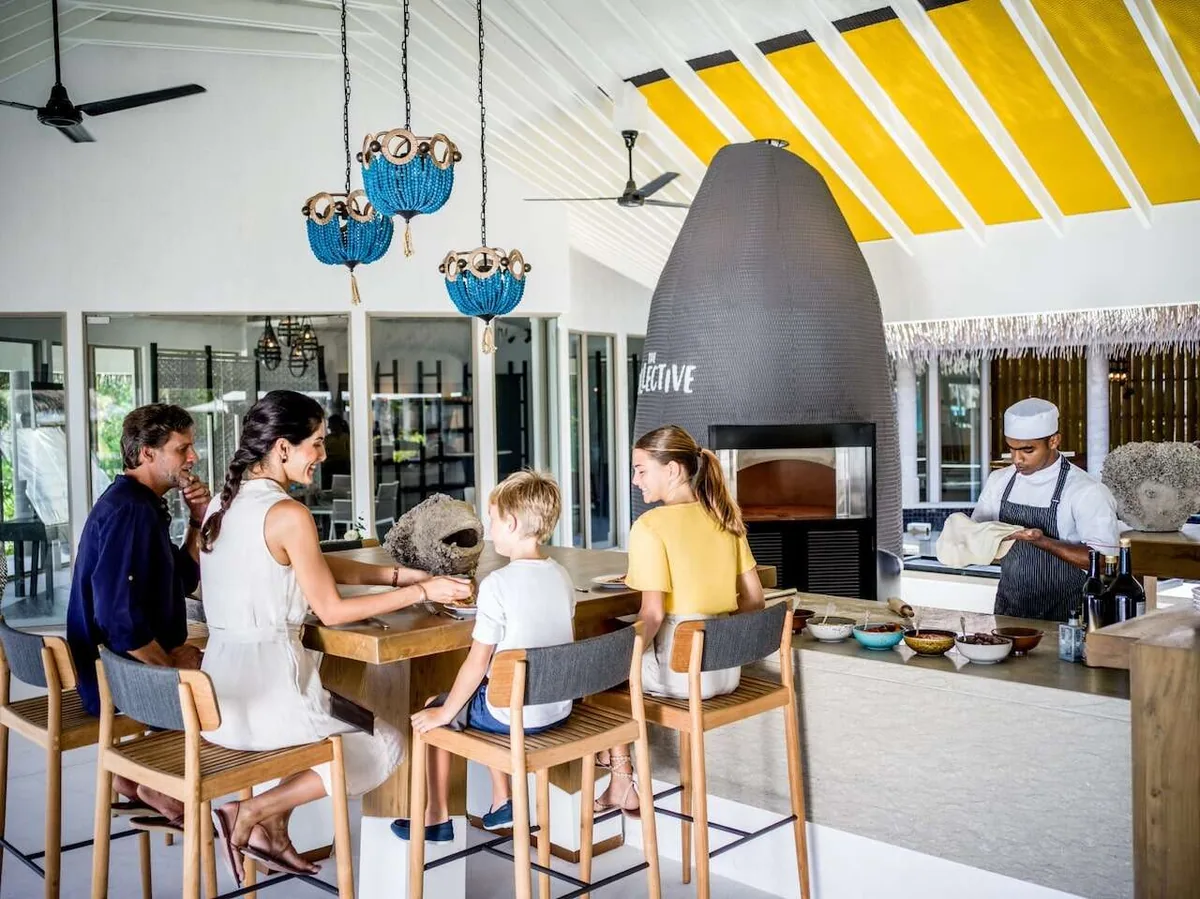InterContinental Maldives Maamunagau Resort, dining, Raa Atoll, Maldives, The Collective family interactive kitchen with the chef demonstrating pizza prep at the marble counter against the matte-black conical pizza oven, family of four (parents and two children) seated at the counter, blue-raffia chandelier pendants under the yellow-striped rafter ceiling.