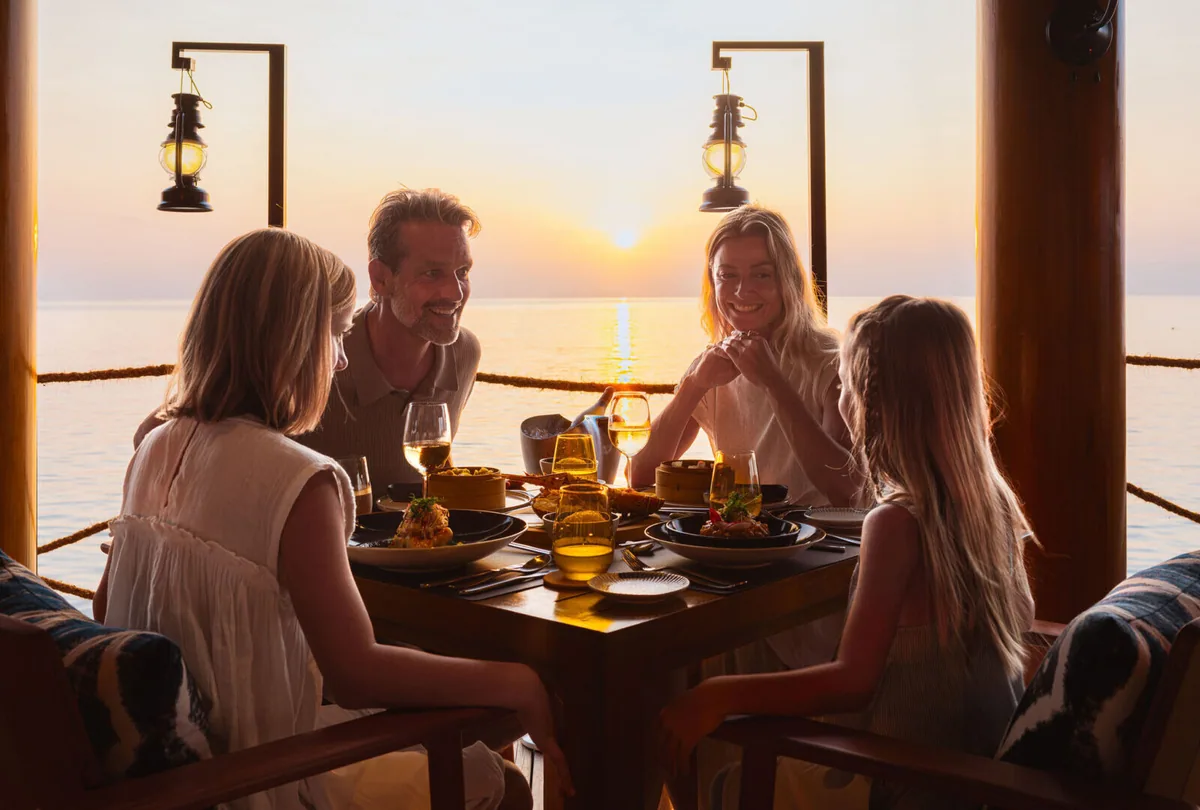 InterContinental Maldives Maamunagau Resort, dining, Raa Atoll, Maldives, Fish Market overwater dining deck at sunset with a multi-generation family of four (parents and two children) at the open table, hurricane-lantern sconces flanking the rope-rail balcony, the lagoon horizon catching the setting sun.