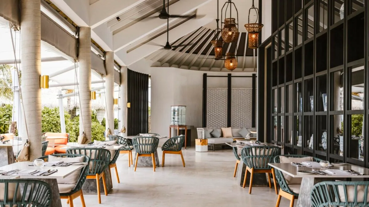 InterContinental Maldives Maamunagau Resort, setting, Raa Atoll, Maldives, The Retreat adults-only sub-property dining pavilion interior with white peaked rafters and ribbed ceiling fans, woven raffia pendant lanterns, saffron-and-orange banquette accents, green woven-back chairs at white tables, vertical screen detailing.