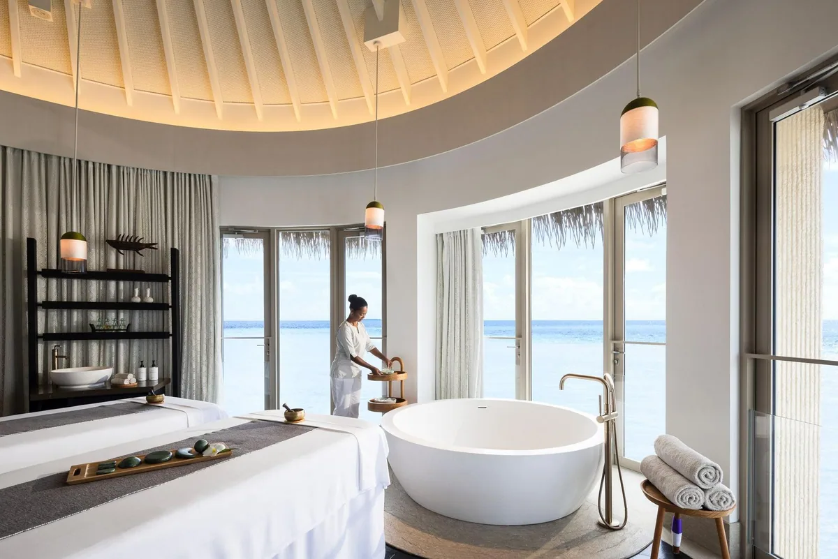 InterContinental Maldives Maamunagau Resort, spa, Raa Atoll, Maldives, AVI SPA overwater treatment pavilion with the signature circular domed ceiling rising above the massage bed and freestanding round soaking tub, the therapist preparing herbs at the side, lagoon visible through full-height windows on both sides.