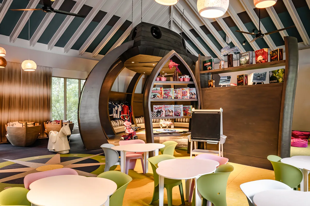 InterContinental Maldives Maamunagau Resort, lifestyle, Raa Atoll, Maldives, Planet Trekkers Kids Club interior with the signature wooden pirate-ship play frame at the centre, ceiling fans under teal-painted rafters, pastel cloud-cutout chair backs around the kids' tables, art easel, bookshelves loaded with board games (Catan, Twister) and craft magazines, kids' clothing rack along the wall.