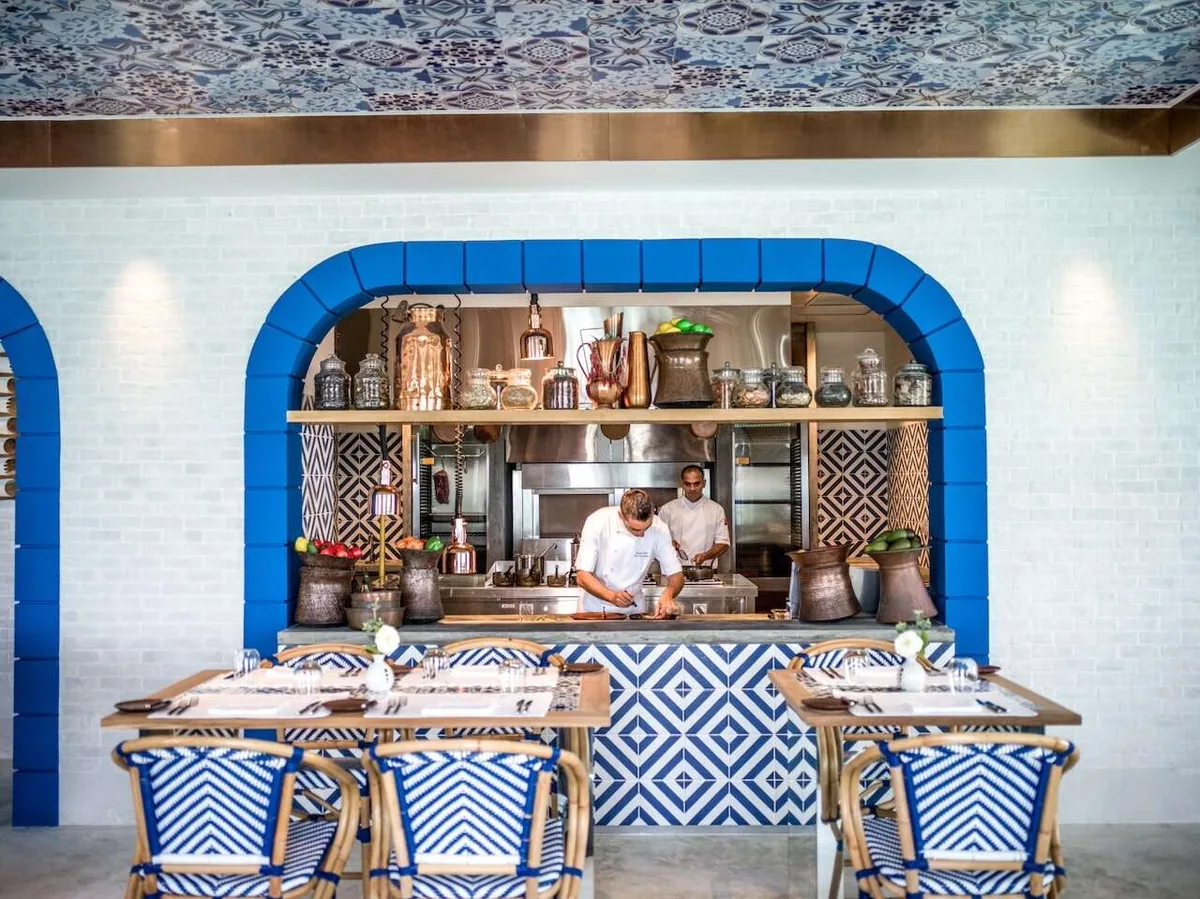 InterContinental Maldives Maamunagau Resort, dining, Raa Atoll, Maldives, The Lighthouse open kitchen with the chefs at the central counter, blue-arched openings framing the kitchen brick wall, intricate blue-and-white Mediterranean-tile floor and bar facing, copper utensils and preserves hung above, woven blue-and-white bistro chairs at the tables.