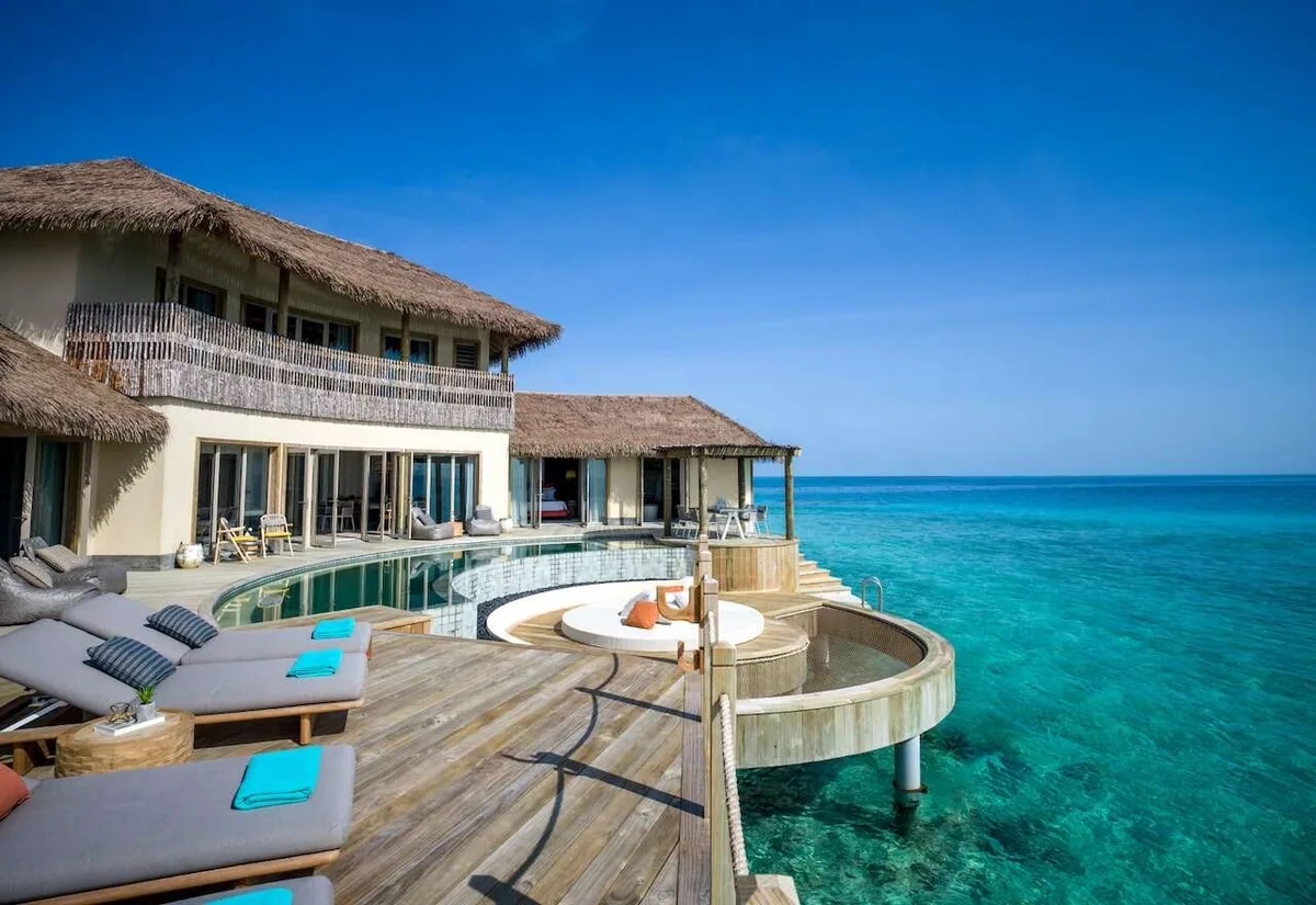 InterContinental Maldives Maamunagau Resort, villa multi bedroom, Raa Atoll, Maldives, 3-Bedroom Overwater Pool Residence exterior, two-story thatched pavilion with wraparound infinity pool, semi-submerged circular glass-bottom relaxation pod above the lagoon, sunken loungers with turquoise towels, wooden deck steps down to the water.