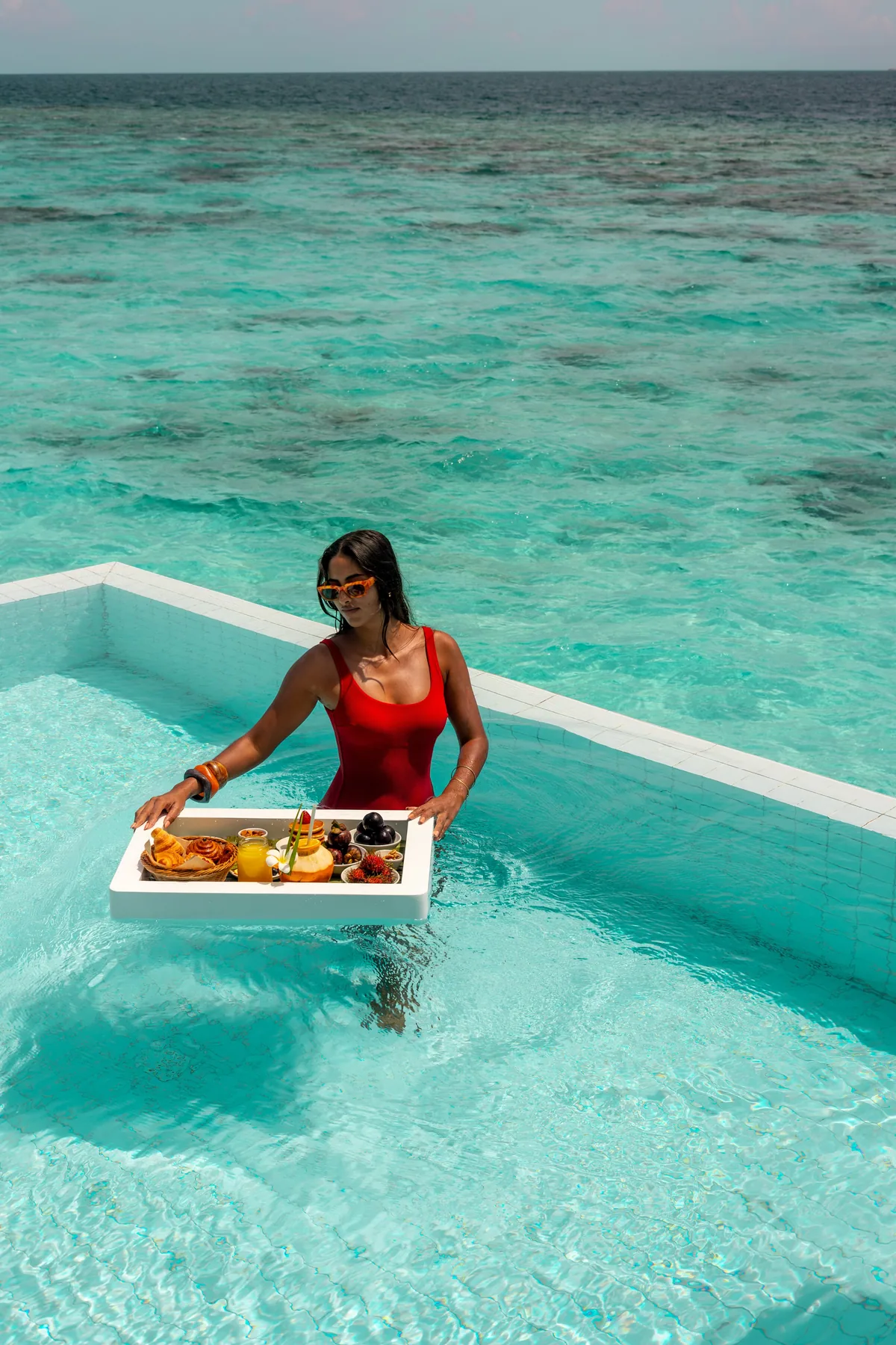 Sun Siyam Iru Veli, dining, Dhaalu Atoll, Maldives, Floating breakfast in the overwater villa plunge pool, a guest in a white-tile infinity-edge pool with a floating tray of pastries, fresh fruit, and juices (the property's signature in-villa dining moment), open-lagoon view stretching to the horizon.