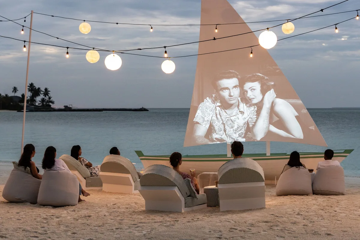 Sun Siyam Iru Veli, activity, Dhaalu Atoll, Maldives, Beach cinema setup at dusk, a classic black-and-white film projected onto the sail of a beached dhoni-style boat, white-sand-set bean bags and canopy chairs facing the screen, string-light overhead with glowing globe pendants, guests gathered for the evening programming.