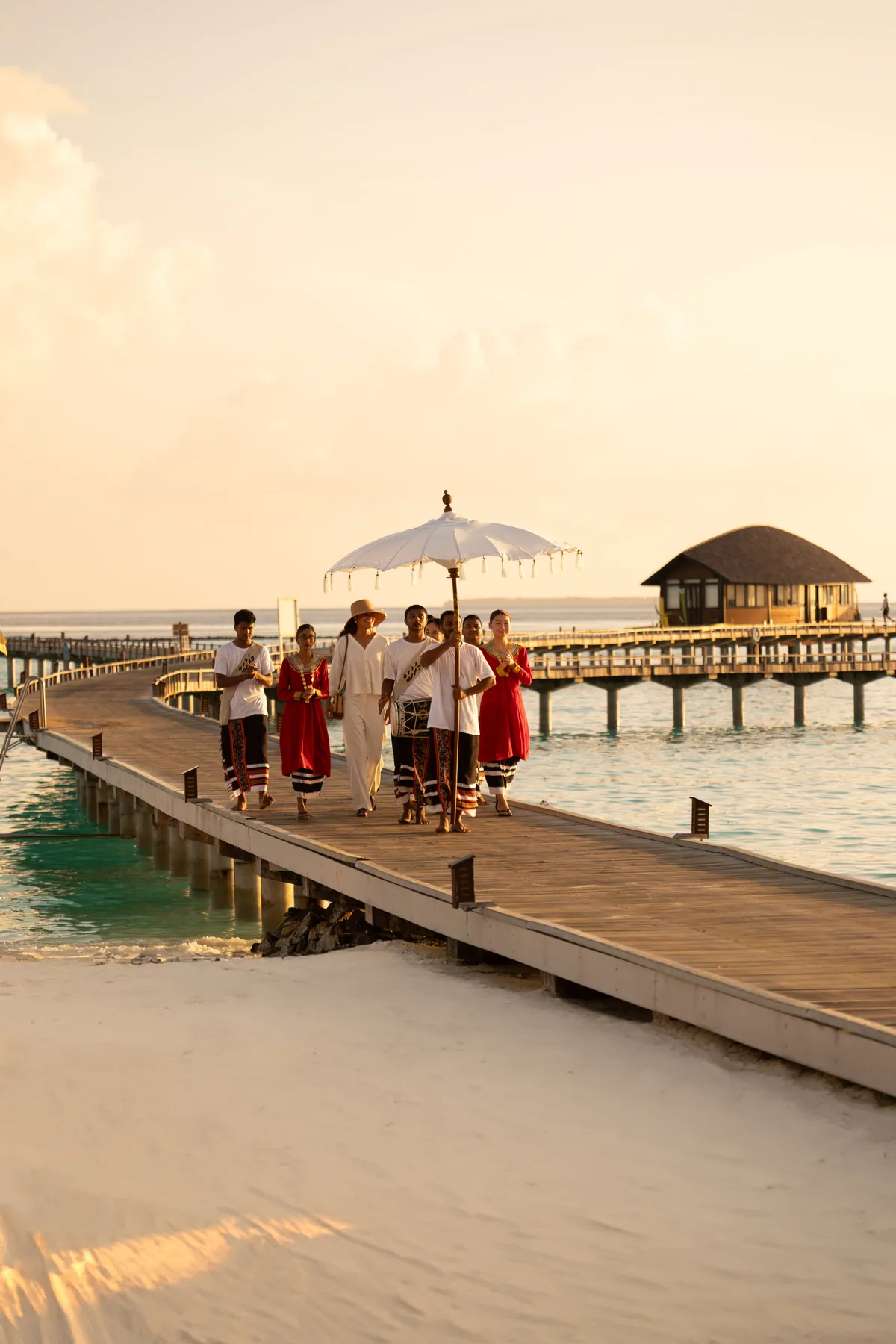 Sun Siyam Iru Veli, setting, Dhaalu Atoll, Maldives, Ceremonial arrival on the jetty walkway at golden hour, a parasol bearer with musicians and welcome dancers in traditional Maldivian dress, the long timber walkway extending across the lagoon with a thatched-roof pavilion at right.