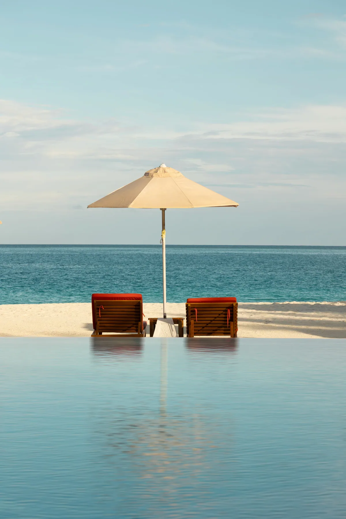 Sun Siyam Iru Veli, setting, Dhaalu Atoll, Maldives, Main pool with timber-frame sun loungers in red cushions under a white parasol, infinity edge reflecting the lagoon, white-sand strip and open horizon beyond.