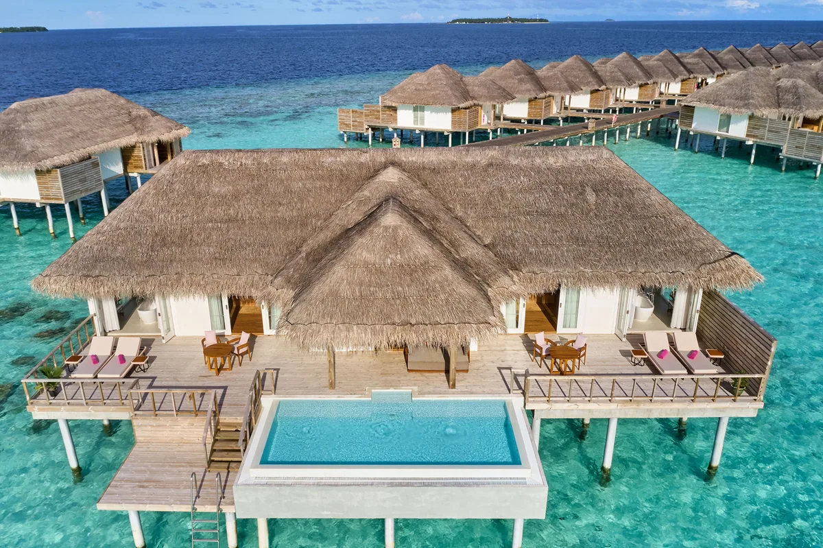 Sun Siyam Iru Veli, villa overwater, Dhaalu Atoll, Maldives, King Ocean Suite from above, a large two-bedroom over-water pavilion with central thatched roof and twin flanking suite wings, central infinity pool, the signature crescent over-water villa row extending in both directions, signature coral-pink loungers (Sun Siyam pop-color) on both decks.