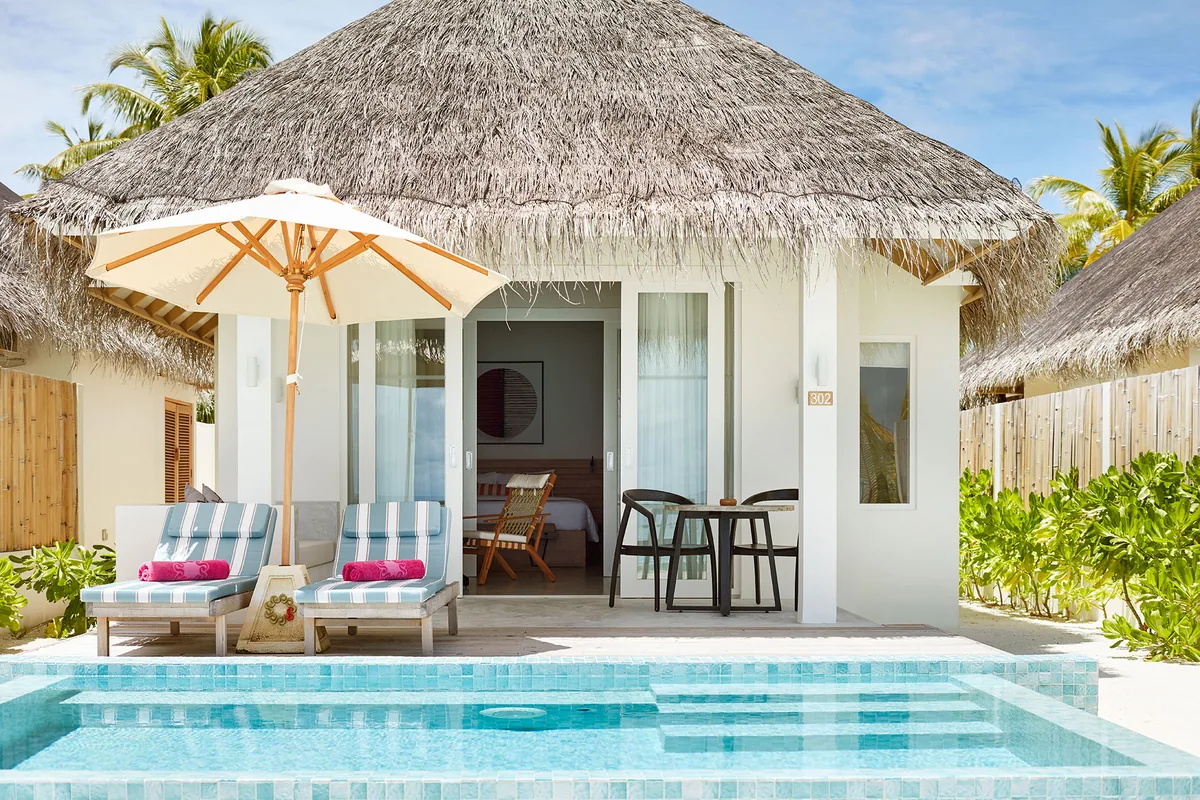 Sun Siyam Iru Veli, villa beach, Dhaalu Atoll, Maldives, Family Suite with Pool exterior, a thatched-roof villa with timber-clad outer walls and a private rectangular pool, sun loungers with the signature pink towels (Sun Siyam pop-color), white umbrella, glass-slider into the villa interior with dining set on the deck.
