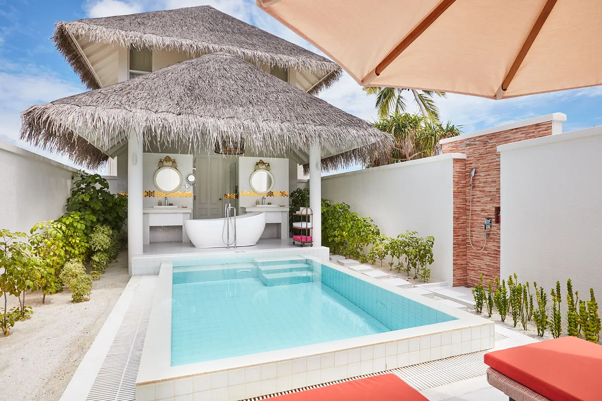 Sun Siyam Iru Veli, villa interior, Dhaalu Atoll, Maldives, Grand Beach Suite open-air bathroom, twin thatched pavilions framing a private dressing-and-bath area with twin round-mirror vanities and a freestanding tub, blue-mosaic plunge pool in the foreground, brick-clad outdoor shower at right, coral pink lounger cushions in the corner.
