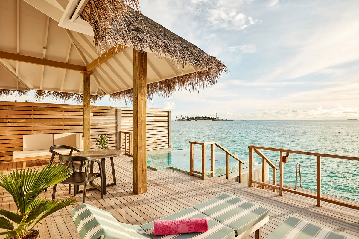 Sun Siyam Iru Veli, villa overwater, Dhaalu Atoll, Maldives, Ocean Suite with Pool overwater deck close-up, the thatched-roof pavilion edge, timber deck with a striped sun lounger and a coral-pink towel (signature Sun Siyam pop-color), private mosaic-edge plunge pool with steps down to the lagoon, slatted-wood privacy screen.