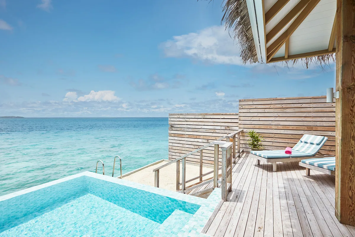 Sun Siyam Iru Veli, villa overwater, Dhaalu Atoll, Maldives, Dolphin Suite overwater deck, a wider view of the timber deck with a mosaic-tile plunge pool extending to an infinity edge over the lagoon, slatted wood privacy screens, striped sun lounger with pineapple welcome.