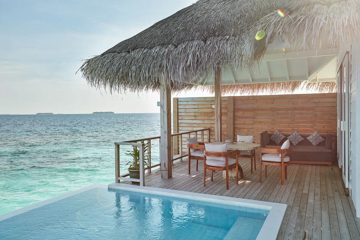 Sun Siyam Iru Veli, villa overwater, Dhaalu Atoll, Maldives, Grand Ocean Suite overwater deck with dining setup, a thatched-eave pavilion framing an outdoor dining set and a grey embroidered-cushion sofa lounge, mosaic-tile plunge pool in the foreground, sun-flare through palm fronds and open-lagoon view beyond.