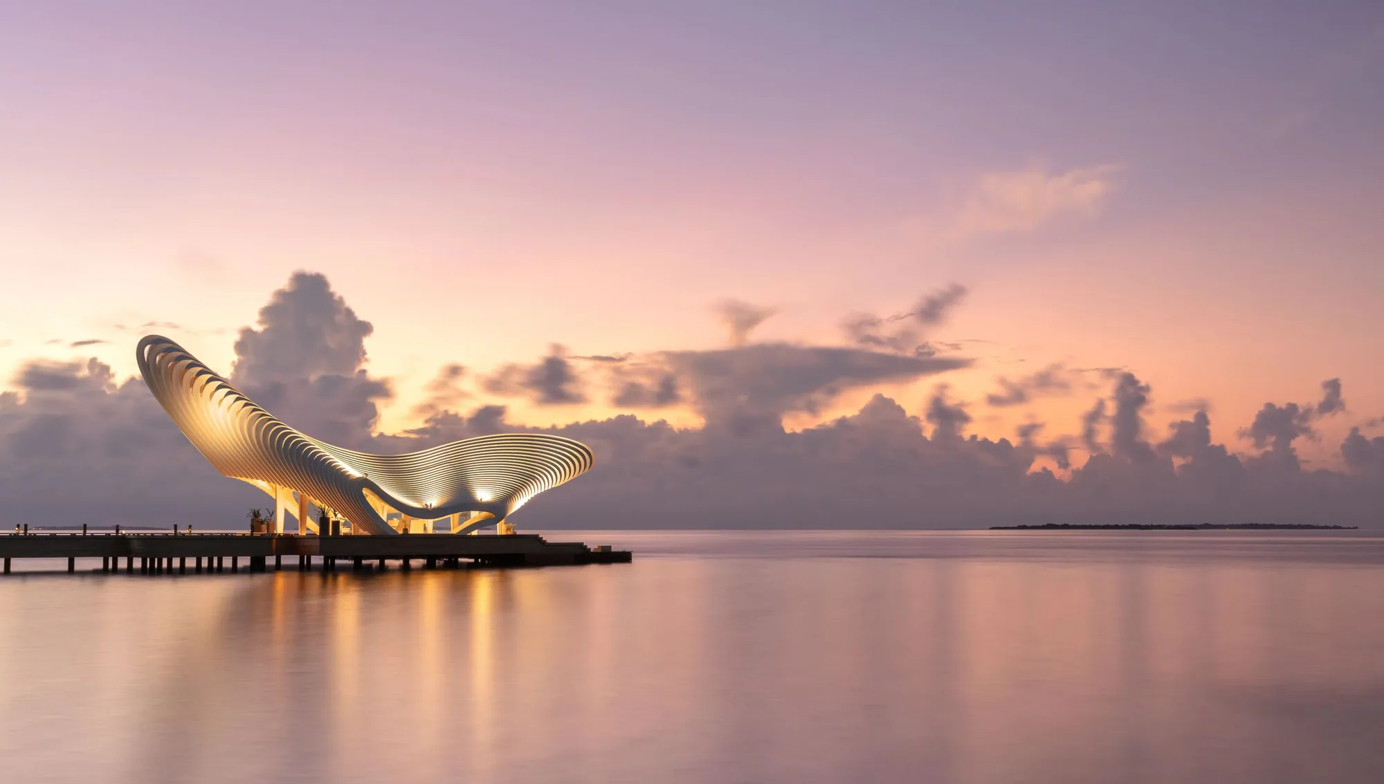 JOALI BEING, arrival, Raa Atoll, Maldives, Gate of Zero, the sculptural ribbed-arch arrival pavilion on the jetty at twilight, illuminated curves reflecting on the glass-calm sea, the property's signature welcome installation.