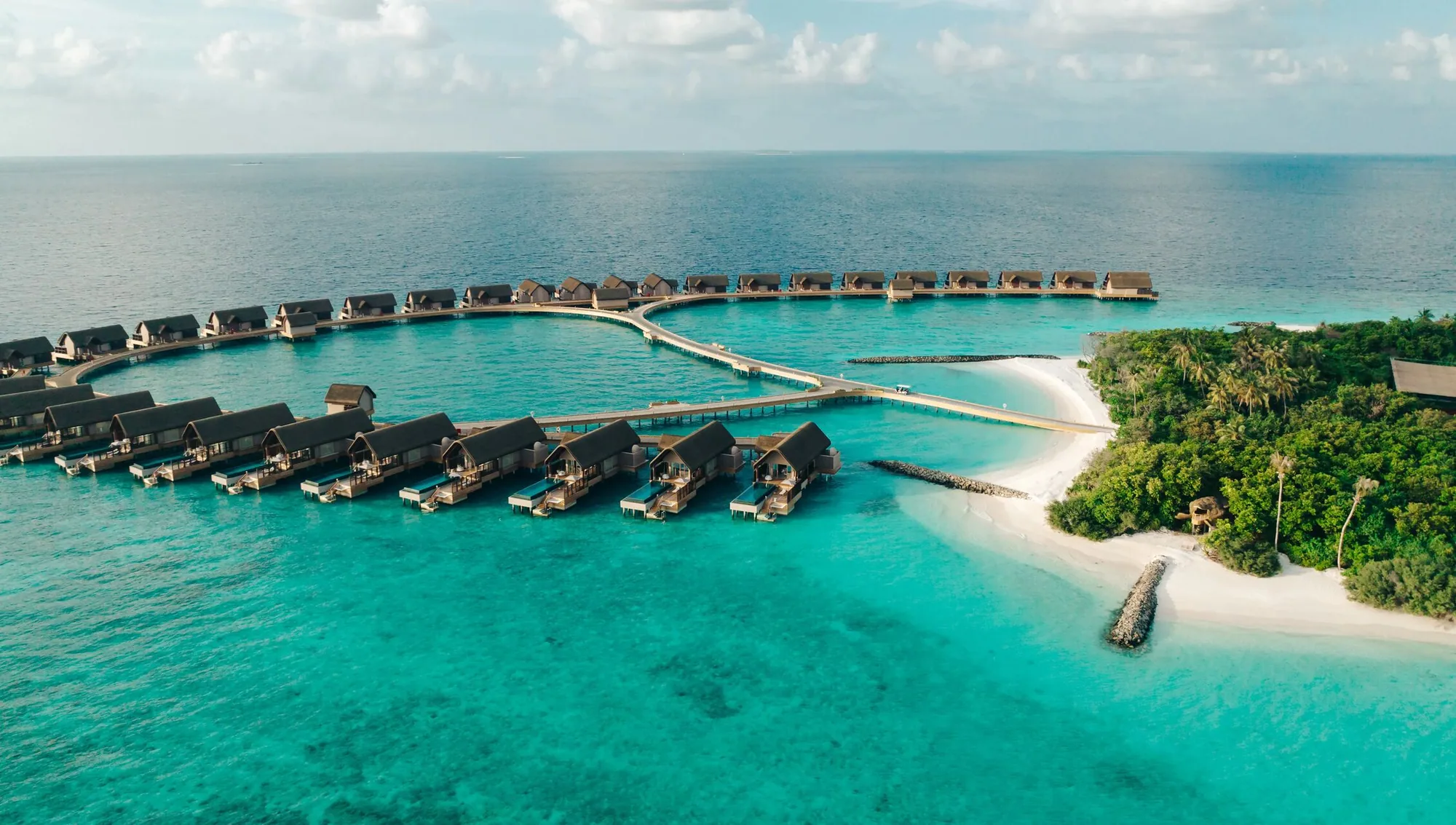 JOALI BEING, setting, Raa Atoll, Maldives, Bodufushi island aerial, curved overwater villa ring tracing the lagoon perimeter, white-sand jetty linking the back-of-house and vegetated island interior.