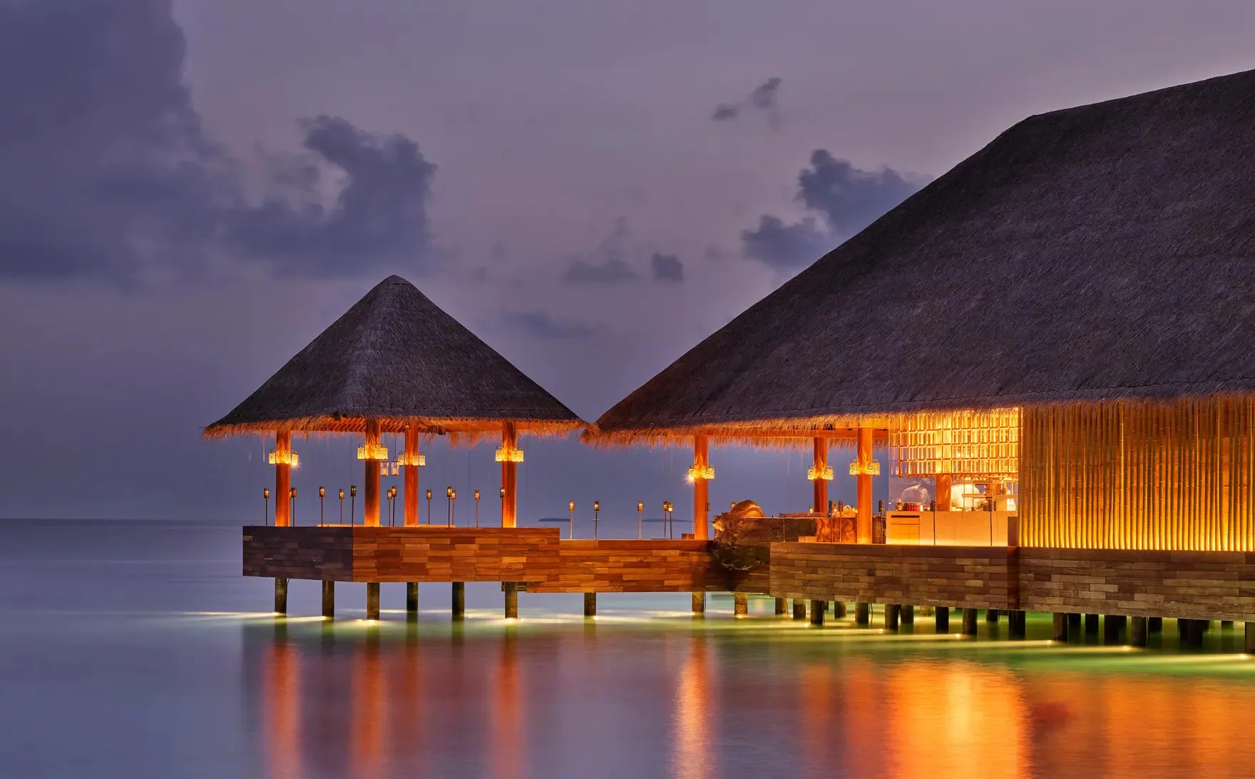 JOALI Maldives, dining saoke, Raa Atoll, Maldives, Saoke at twilight: twin pyramid-thatched overwater pavilions glowing from within, the Japanese teppanyaki counter on stilts over the lagoon, deep blue dusk sky with warm reflections on the water.