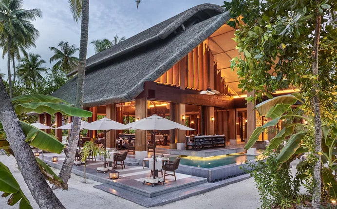 JOALI Maldives, dining vandhoo, Raa Atoll, Maldives, Vandhoo, the all-day pavilion: the property's iconic curved origami-thatched roof, warm timber colonnade, open-air dining terrace with white parasols, lily-pad reflection pool front, dense palm framing.