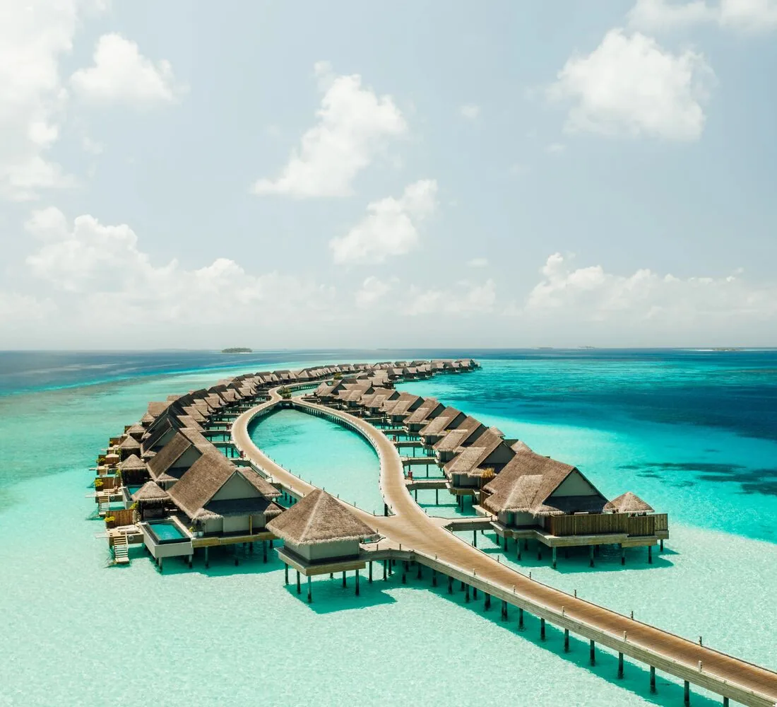 JOALI Maldives, setting, Raa Atoll, Maldives, JOALI's signature horseshoe-shaped overwater boardwalk arc seen from the air: twin rows of thatched-pyramid villas curving around a central oval lagoon, a single observation pavilion at the apex, the timber walkway connecting back to Muravandhoo across the turquoise lagoon.