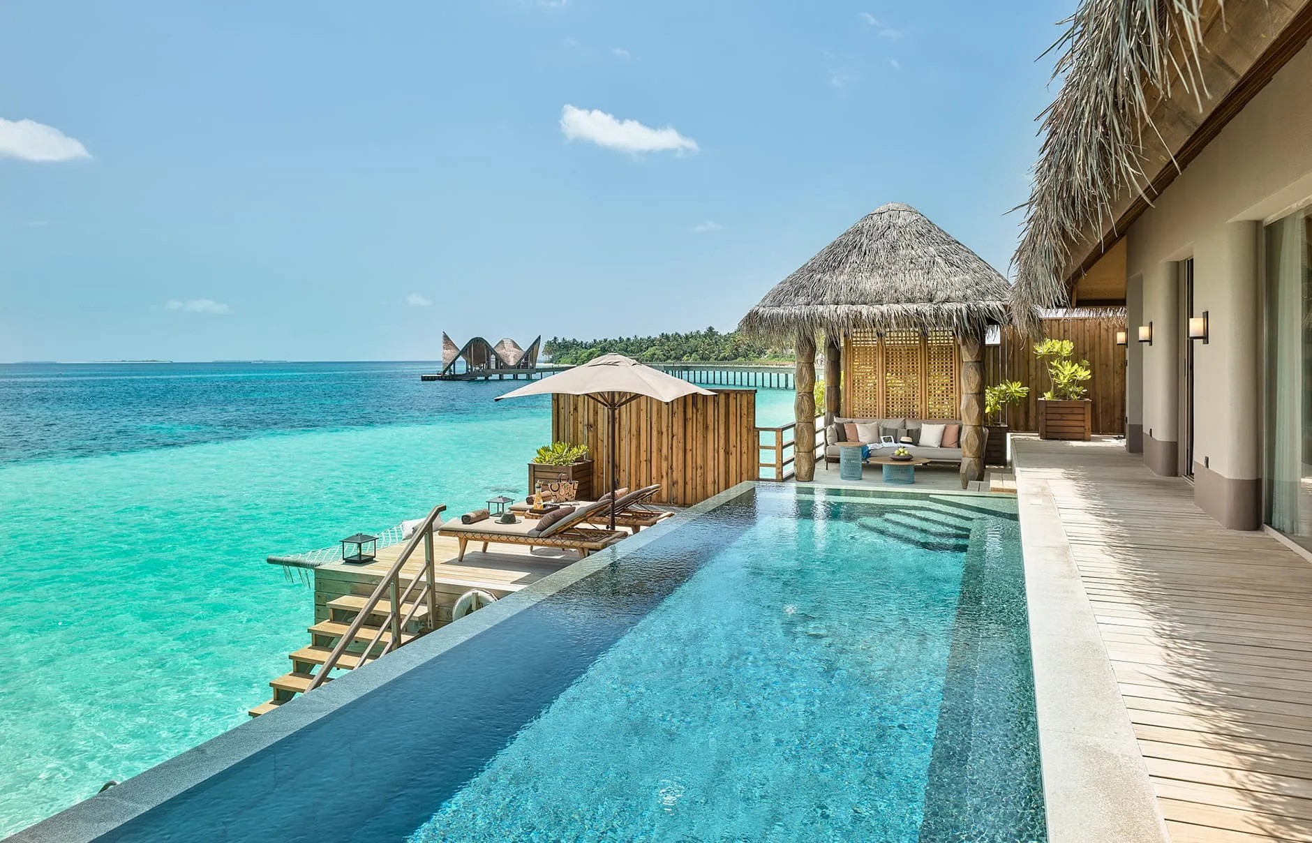 JOALI Maldives, villa overwater, Raa Atoll, Maldives, Sunset Water Villa with Pool: an infinity-edge plunge pool running off the teak deck toward the horizon, twin sun loungers, a thatched pavilion with twin daybeds, a neighbouring villa pavilion just visible at the deck edge.