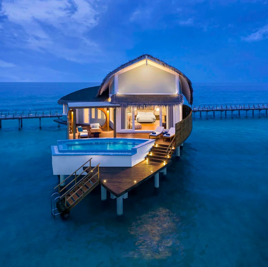 JW Marriott Maldives Resort & Spa, Overwater Pool Villa twilight exterior with scalloped thatched roof and lit pool, Shaviyani Atoll, Maldives