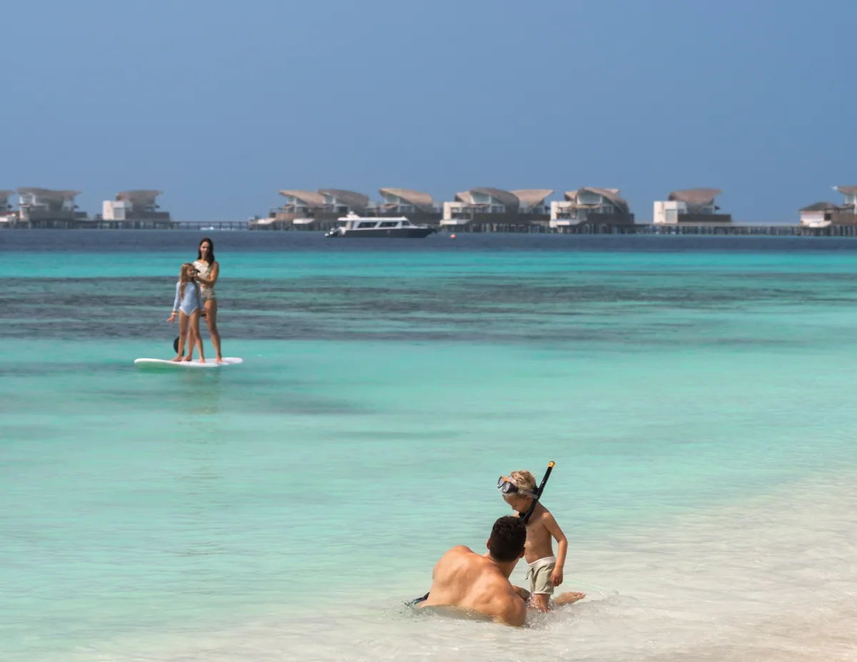 JW Marriott Maldives Resort & Spa, family on property beach with OWV cluster anchoring the horizon, Shaviyani Atoll, Maldives