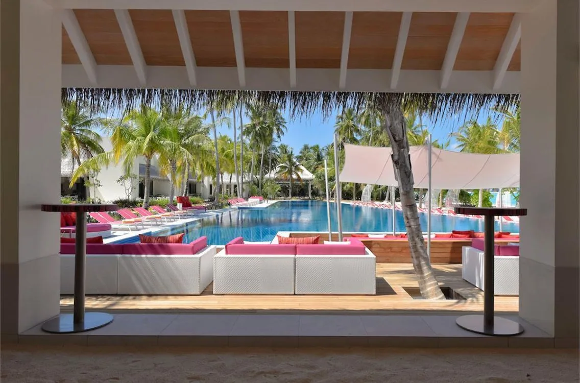 Kandima Maldives, dining, Dhaalu Atoll, Maldives, Pool lounge framed by a thatched-roof architectural opening, white modular sofas with signature pink cushions, bar tables, palms and a sail-cloth shade, the long pool stretching out with sun loungers along the edge.