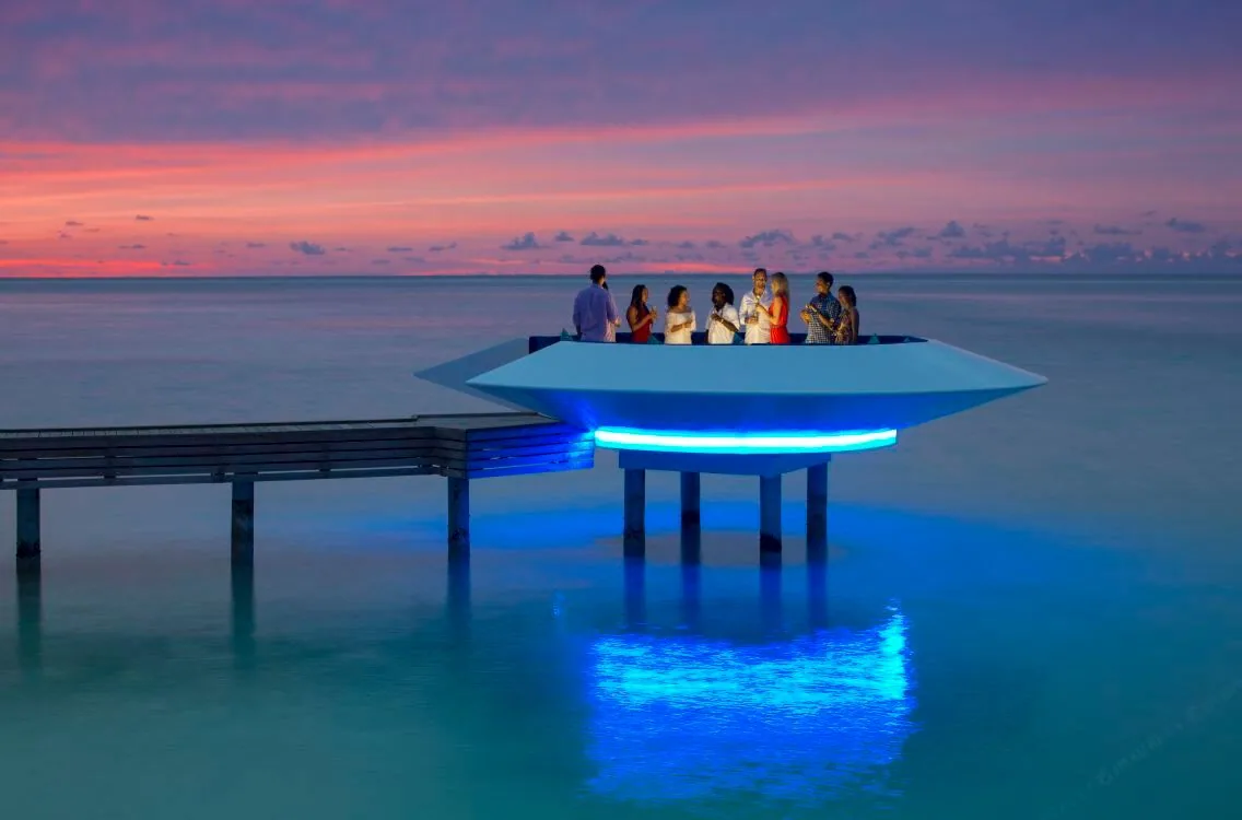 Kandima Maldives, dining, Dhaalu Atoll, Maldives, Signature overwater UFO-shape sundowner platform with neon-blue underlighting at dusk, group of guests with sunset cocktails on the floating disc, pink-orange sunset horizon, blue-neon reflection in the lagoon below (the property's signature sundowner spot).