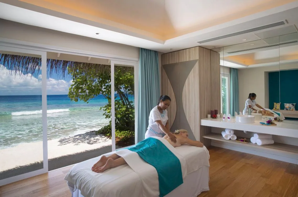 Kandima Maldives, spa, Dhaalu Atoll, Maldives, eSKAPE Spa treatment room, a therapist working on a guest at the massage table, glass sliding doors opening to the beach and ocean view, signature aqua-teal towel and curtains (pop-color), light hardwood floor and pale cabinetry.