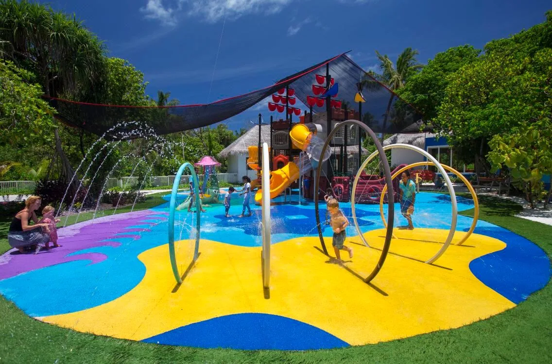 Kandima Maldives, activity, Dhaalu Atoll, Maldives, Kids' splash pad, colorful pop-color play structure with water jets and spray hoops, children playing on the painted yellow-and-purple-and-blue rubber floor, pirate-ship themed slide visible in the background.
