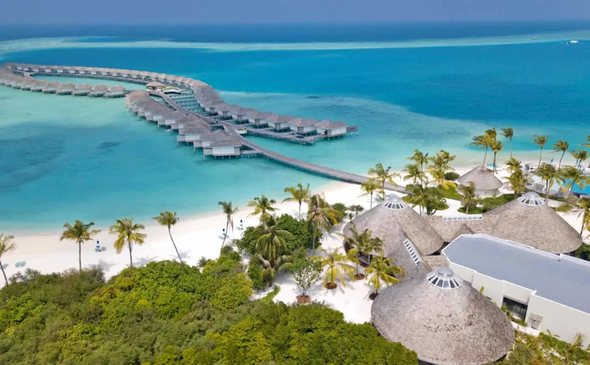 Kandima Maldives, aerial, Dhaalu Atoll, Maldives, Mid-spine aerial showing the connection between the main-island activity cluster (round-thatched-roof dining pavilions in foreground) and the over-water villa row across the lagoon, white-sand beach and palm canopy interior, distant island on horizon.