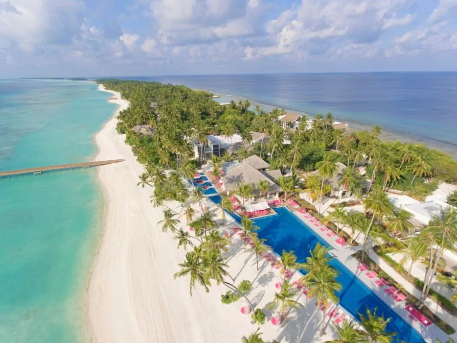Kandima Maldives, aerial, Dhaalu Atoll, Maldives, Aerial of the property's signature 100-metre main pool (one of the country's longest resort pools), red sun loungers and red umbrellas along both edges, thatched pavilions and palm canopy framing, beach front continuing along the long-axis island.