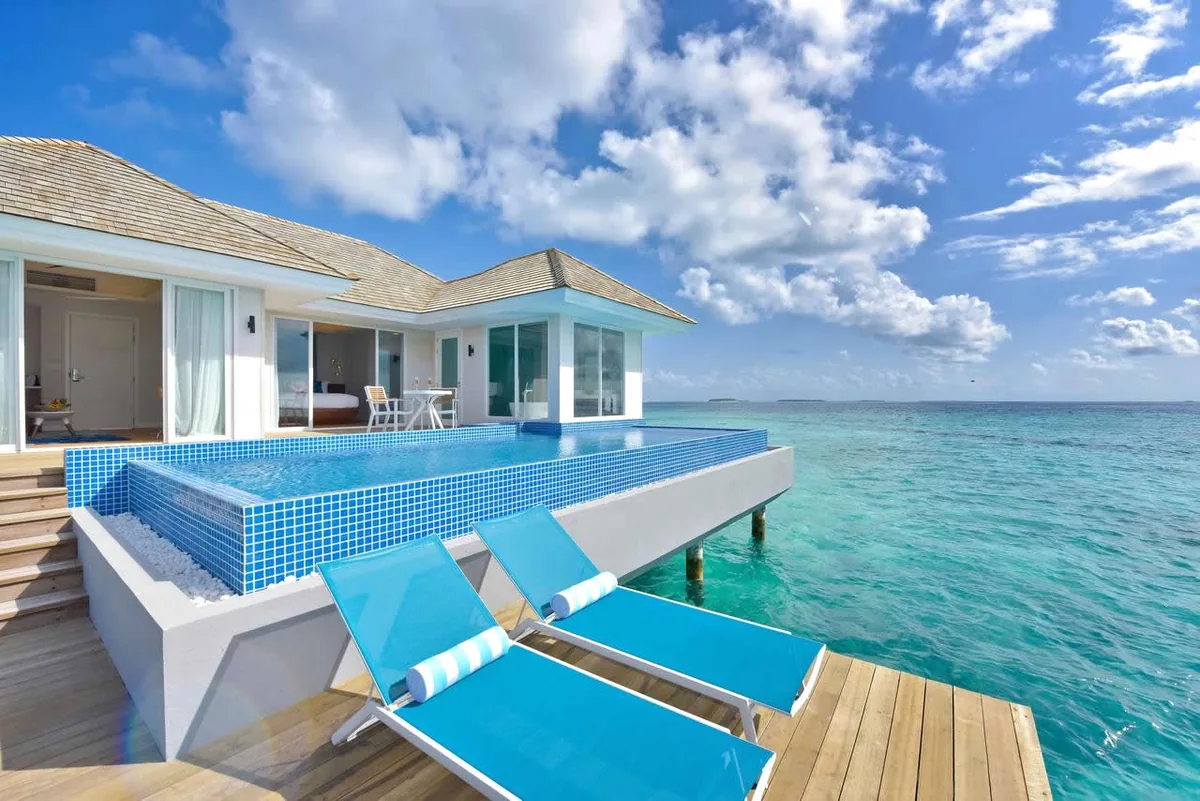 Kandima Maldives, villa overwater, Dhaalu Atoll, Maldives, Aqua Villa with Pool exterior, a single-storey overwater pavilion with shingle roof, signature aqua-mosaic-tile infinity pool (the Kandima pop-color signature) flush with the lagoon, aqua-blue sun loungers on the timber deck, glass-walled bedroom and open-ocean view.