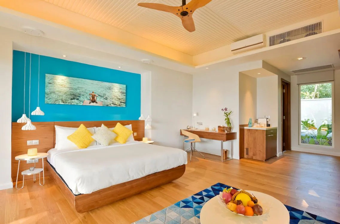 Kandima Maldives, villa interior, Dhaalu Atoll, Maldives, Villa bedroom interior, king bed with hardwood frame, signature aqua feature wall behind a coral-reef long-form photo art (Kandima pop-color palette), yellow throw pillows on white linens, wood floor and ceiling fan, fruit bowl on a geometric blue floor rug.