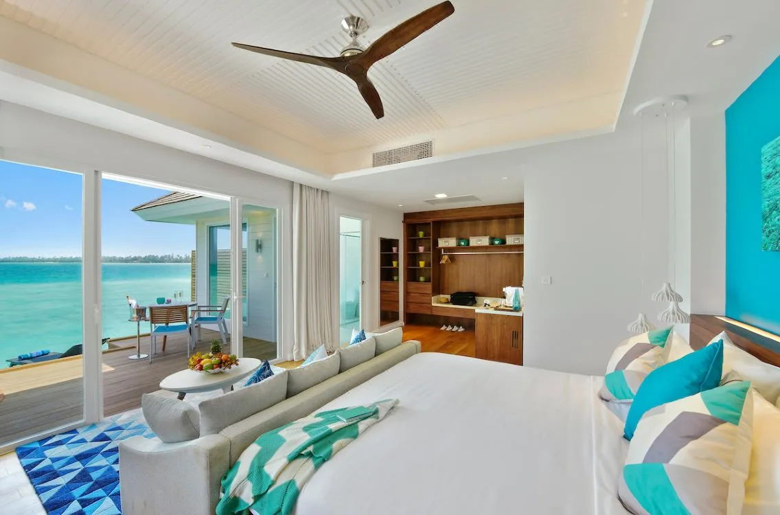 Kandima Maldives, villa interior, Dhaalu Atoll, Maldives, Aqua Villa overwater bedroom, king bed with chevron throw and aqua and grey geometric pillows (signature palette), wood feature wall with display shelving, ceiling fan, glass doors opening to the overwater deck with breakfast setup and lagoon view at left.
