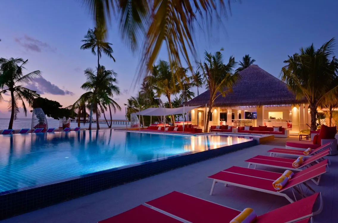 Kandima Maldives, dining, Dhaalu Atoll, Maldives, Beach Club at dusk, signature red sun loungers along the infinity pool edge, thatched-roof bar pavilion lit from within, palm canopy at the perimeter, lavender twilight sky and distant lagoon.